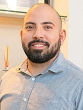 Homem sorridente com barba, usando camisa social cinza clara, posando em ambiente corporativo iluminado.
